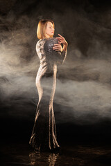 Woman in a shiny silver dress moves and dances on a black background and smoke