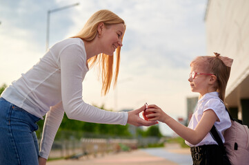 woman giving apple to schoolgirl. outdoor interaction highlights generosity and education bonding. smiling child with backpack, casual urban background creates warm, inviting scene.