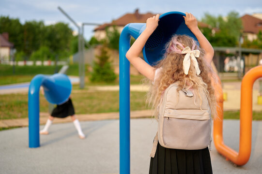 children playing with interactive playground equipment in sunny park. girl with backpack listens through colorful sound tubes, engaging in outdoor learning and communication activity