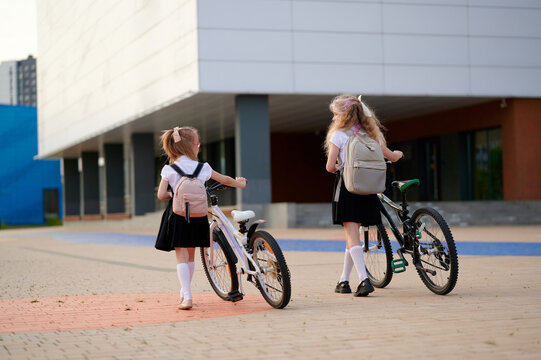 two schoolgirls with bicycles walking to school building. backpacks, uniforms, and bicycles highlight morning routine, education, and healthy lifestyle. modern architecture in background - Powered by Adobe