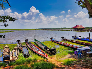 Colorful traditional fishing boats and tourist boats, and a long pier, on the shore of Thale Noi (Thalay Noi), a protected wetland and bird sanctuary in Phatthalung Province in Southern Thailand