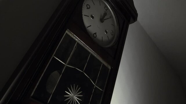 Dramatic low-angle, Dutch tilt zoom-in on a vintage pendulum wall clock. The quick movement of the hands suggests a time-lapse, symbolizing the passage of time. The moody, low-key lighting