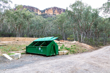 Photograph of a large green steel industrial skip rubbish bin with lids open left on the side of a dirt road in a regional forest. 