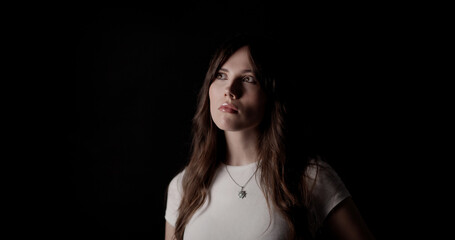 Studio portrait of a young woman with long brown hair wearing a white t shirt and a necklace, looking up on a black background