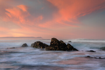 View of dramatic dark rocks rise from the blurred, surging ocean under a vibrant and colorful sunset sky, creating a serene coastal scene, Hermanus, Western Cape, South Africa.