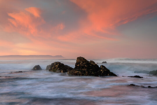 View of dramatic dark rocks rise from the blurred, surging ocean under a vibrant and colorful sunset sky, creating a serene coastal scene, Hermanus, Western Cape, South Africa.