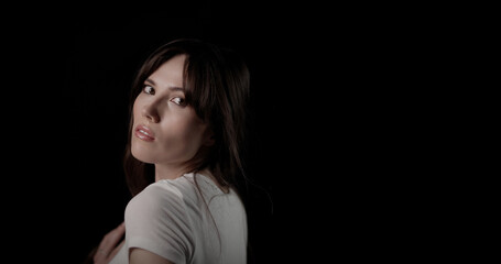 Naklejka premium Portrait of a beautiful young woman with long brown hair, wearing a white t shirt, turning her head and looking back over her shoulder against a black background