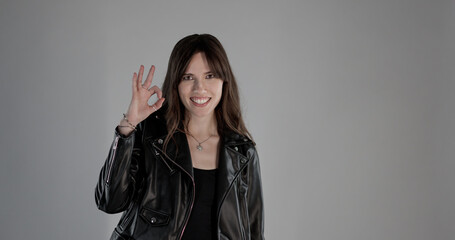 Young woman with leather jacket making ok sign with fingers and smiling on gray background