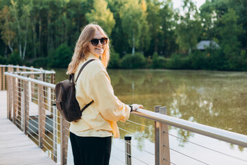 A young happy blonde woman standing on a bridge near a lake on a sunny day. Beautiful morning relax