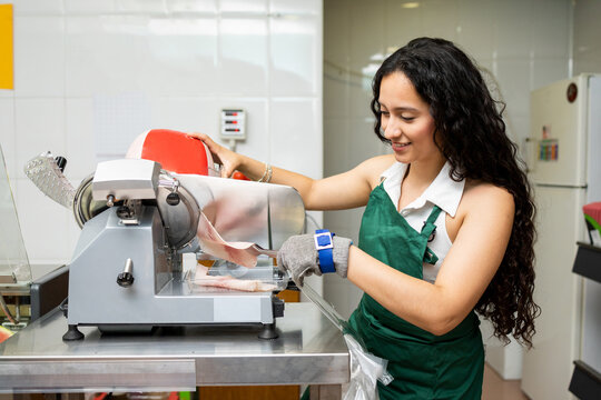 A Latina woman in her 20s and 30s is selling in a small grocery store.The girl is cutting sausage with a slicer.