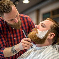 A barber trims the beard of a man in a barbershop. The barber has a short beard and wears a plaid shirt. The client has a long red beard and is seated.
