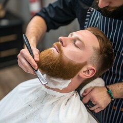 A young Caucasian man with a red beard is receiving a shave at a barbershop. A barber is using a straight razor to trim his facial hair.