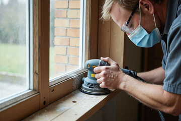 Meticulous worker in a protective face mask uses an orbital sander to smooth an old wooden window frame during home renovation, showcasing detailed craftsmanship, repair, and restoration work