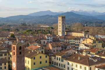 Panoramic view from Guinigi Tower of Basilica di San Frediano and bell tower in Lucca, Italy, with tiled rooftops and mountains.