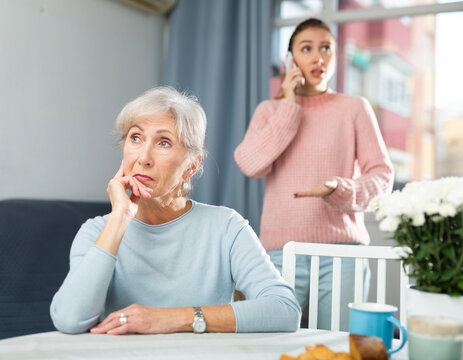 Offended senior woman sitting at table at home and listening her daughter swearing while talking on phone.