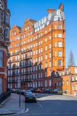 Bright sunshine illuminates the striking red brick building in Kensington, London. The architectural style showcases intricate details typical of the area's historic charm.