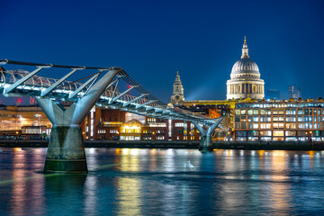As evening falls, the Millenium Bridge elegantly spans the River Thames, showcasing stunning reflections while St. Paul's Cathedral stands majestically illuminated in the background.