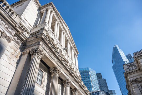 The historic Bank of England building stands prominently against a clear blue sky. Modern skyscrapers surround this iconic structure, showcasing London's architectural blend of old and new.
