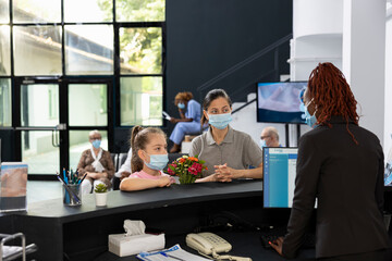 Receptionist greeting mother and child at the hospital front desk, assisting them with registration procedures and safety hygiene policy with the face mask. Cute girl visiting with flowers.