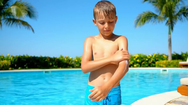Young boy applying sunscreen to his arm next to a swimming pool with palm trees, emphasizing sun protection during summer outdoor activities