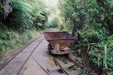An old rusty mine cart sits on tracks in Karangahake Gorge, New Zealand. The tracks lead into a...