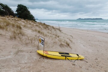 A yellow lifeguard rescue board sits on a sandy beach next to a warning sign about strong rip...