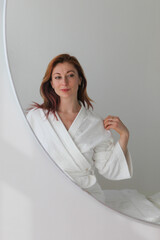Ginger-haired woman, 30s, sitting on massage bed in a spa, wearing a white robe. Looking...