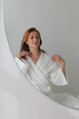 Ginger-haired woman, 30s, sitting on massage bed in a spa, wearing a white robe. Looking...