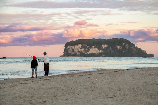 A father teaches his son how to fish on a sandy beach at sunset in Whangamata, Coromandel Peninsula, New Zealand. They are bonding and enjoying quality time together. - Powered by Adobe