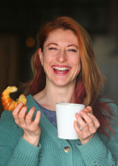 Happy redhead woman enjoying coffee and croissants in a cozy indoor setting.