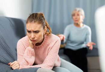 Frustrated young girl sitting on sofa at home while agitated elderly mother reprimanding her....