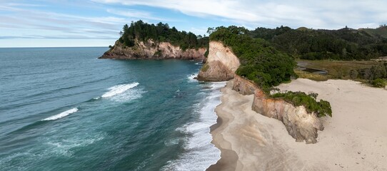 Aerial view of a secluded beach with dramatic cliffs and lush vegetation in Whiritoa, Coromandel Peninsula, New Zealand. The turquoise water meets the sandy shore.