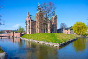 Fototapeta premium Rosenborg Castle showcases its stunning renaissance architecture in Copenhagen, surrounded by tranquil waters and lush greenery on a bright, clear day with blue skies.