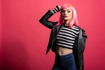 Confident female model posing with arm up wearing bold fashionable clothes against pink background. Stylish attractive woman stands tall with focused eyes and firm body pose, looking at camera.