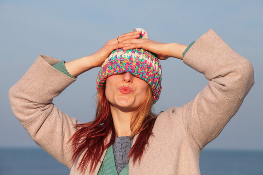 Playful ginger-haired woman, 30s, outdoors by sea, in a colorful outfit and fun hat. Laughing and being silly,. Bright, cheerful spring or fall vibe.