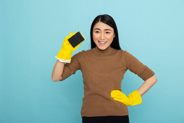 Smiling asian housemaid stands with hand on waist and another holding cleaning sponge. Confident...
