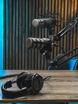 A professional podcast setup features two microphones mounted on adjustable arms, with a pair of headphones resting on the wooden table. The backdrop has a warm, inviting glow.