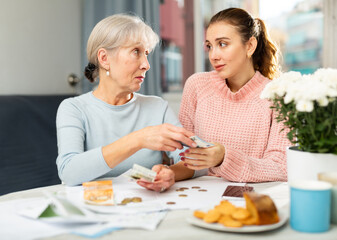 Grandmother sitting at table and giving banknotes to her granddaughter while counting family budget.