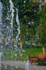 Close-up of fountain water droplets and splashes in an urban park, set against green foliage and a wooden bench, capturing refreshing summer coolness. Photo