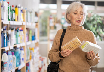 Attentive old woman reviewing packages of clothespins to buy in household goods shop