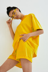 young woman wearing bright yellow oversized t-shirt and shorts with matching yellow sunglasses posing against white background, casual summer fashion style