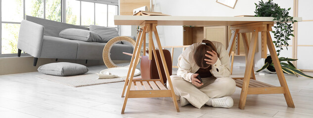 Young woman hiding under desk in living room during earthquake