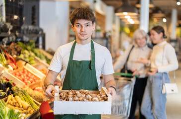 Guy store employee carries box of mushrooms into sales area. Salesperson replenishes stock of goods...