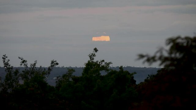Video of the full moon rising moving through the gray clouds of a dramatic sky, birds flying past in the distance.