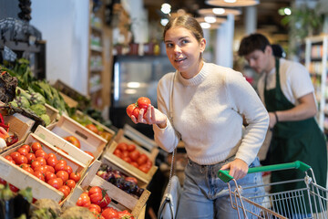 In cozy self-service store, girl customer takes tomatoes out of box. Store visitor chooses best...