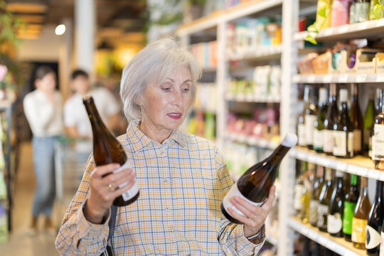 Mature woman examines bottles of wine near display case, reads product label information. Client compares different options for alcoholic drink, chooses bottle of pink wine.