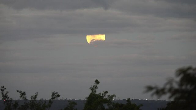 Video of the full moon rising moving through the gray clouds of a dramatic sky, birds flying past in the distance.