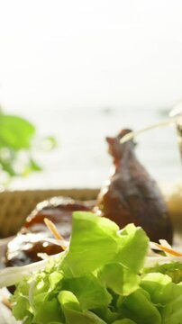 Vertical video. Grilled chicken leg with papaya salad and cold lemonade on rattan tray with sea in background, close up, Traditional Thai Food Concept