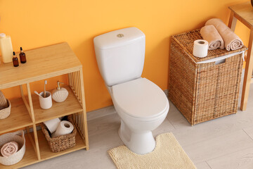 Interior of restroom with toilet bowl, shelving unit and basket near orange wall