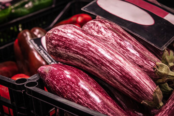 A plastic crate filled with elongated purple and white striped graffiti eggplants, also called Sicilian eggplants, sits among other vegetables at an indoor produce stall in Valencia market, Spain.